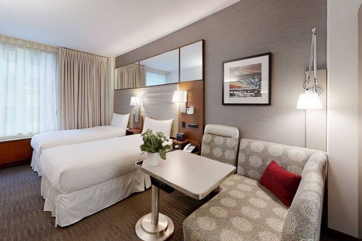 Une chambre d’hôtel avec deux lits, un canapé, une petite table avec une plante, une lampe et un tableau encadré au mur.