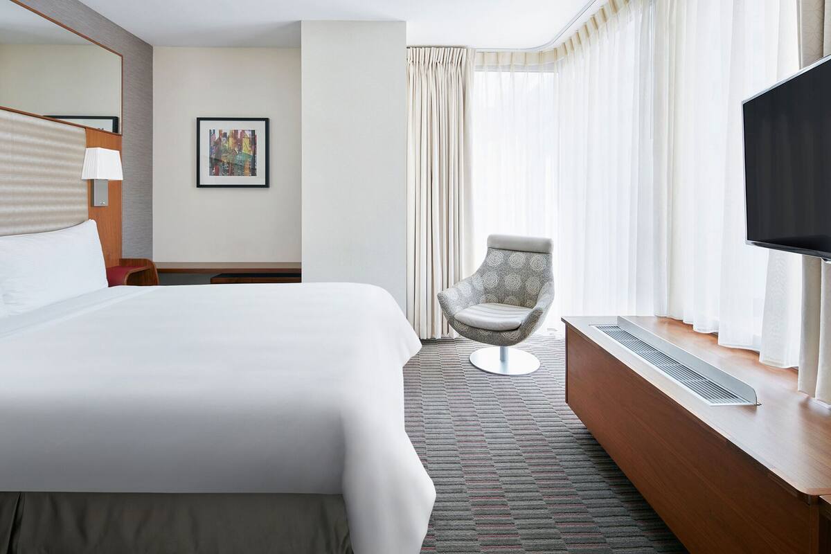 Une chambre d’hôtel avec un grand lit, une chaise, une télévision et une fenêtre avec des rideaux.