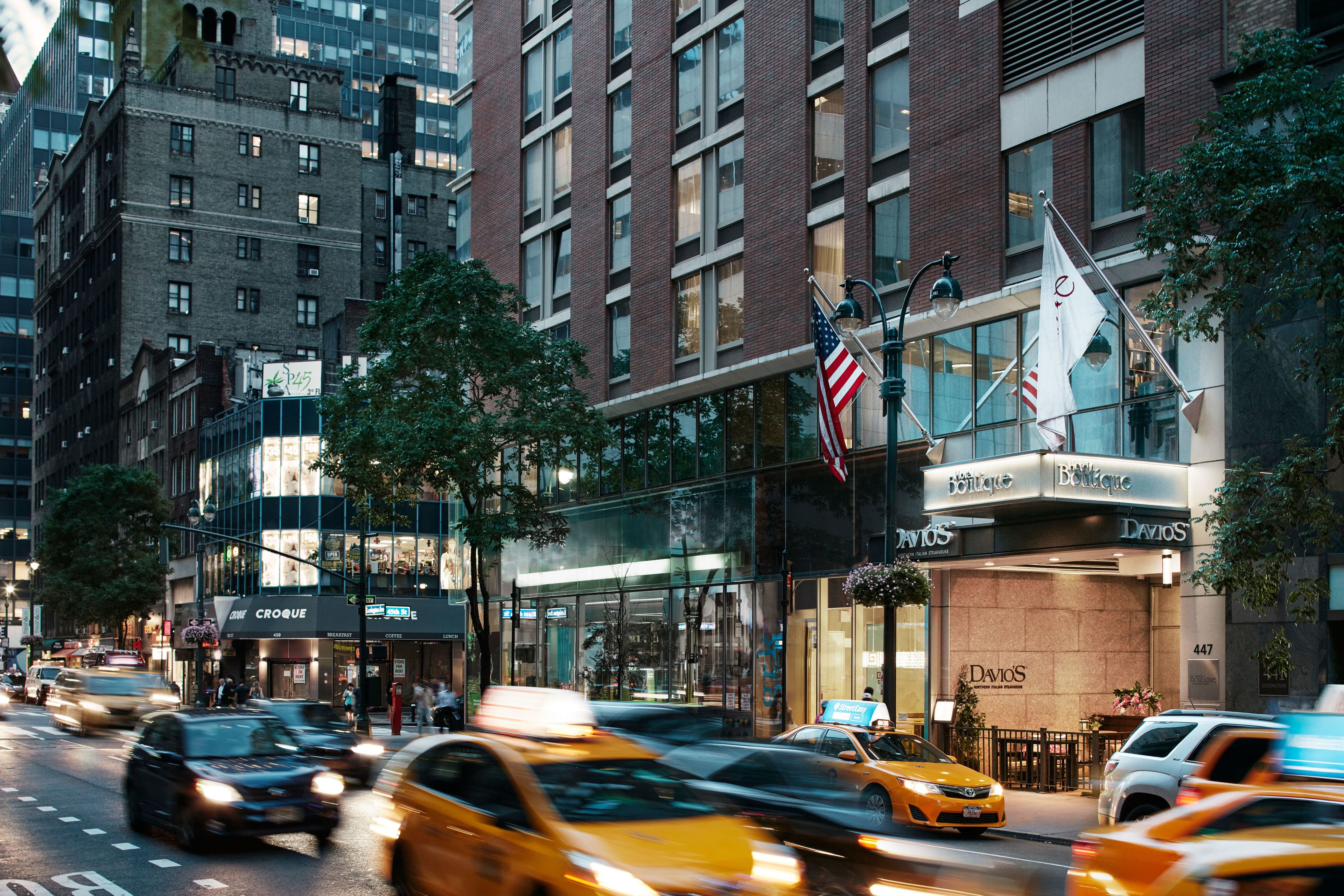 A busy urban street with modern buildings, including a boutique hotel with a glass facade and outdoor seating.