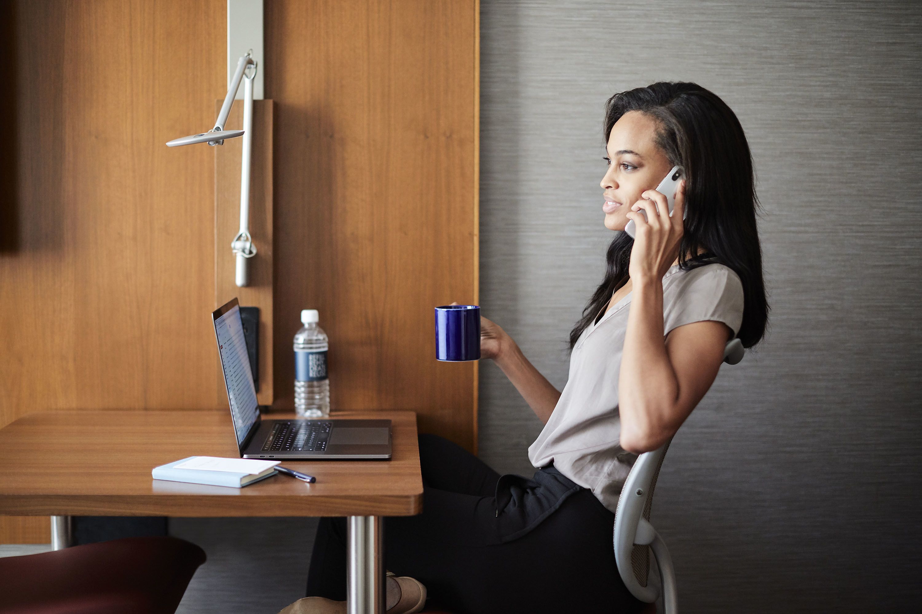 Une personne assise à un bureau en bois, devant un ordinateur portable, avec un carnet et une bouteille d’eau.