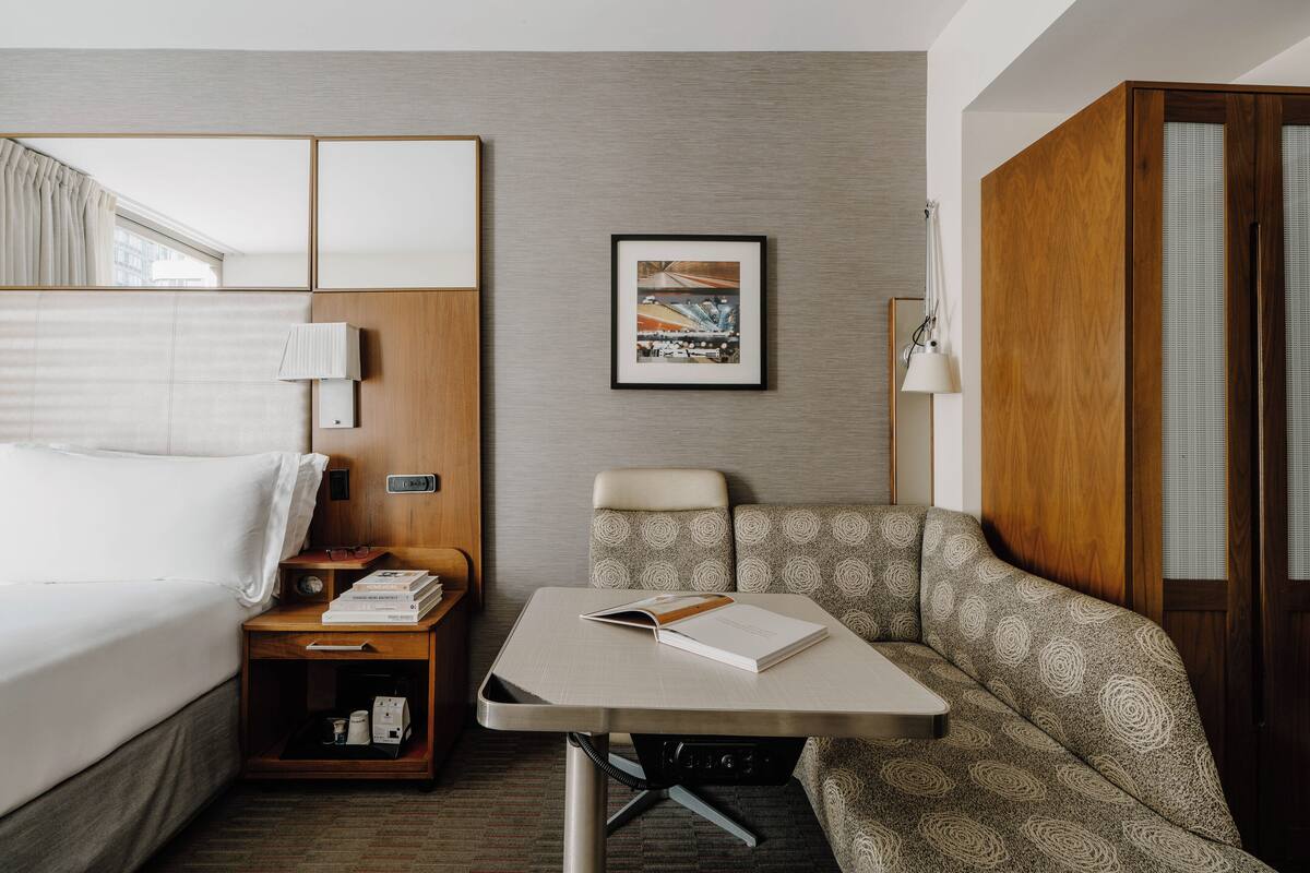 Une chambre d’hôtel avec un lit, une petite table sur laquelle se trouve un livre, une chaise et un tableau encadré accroché au mur.