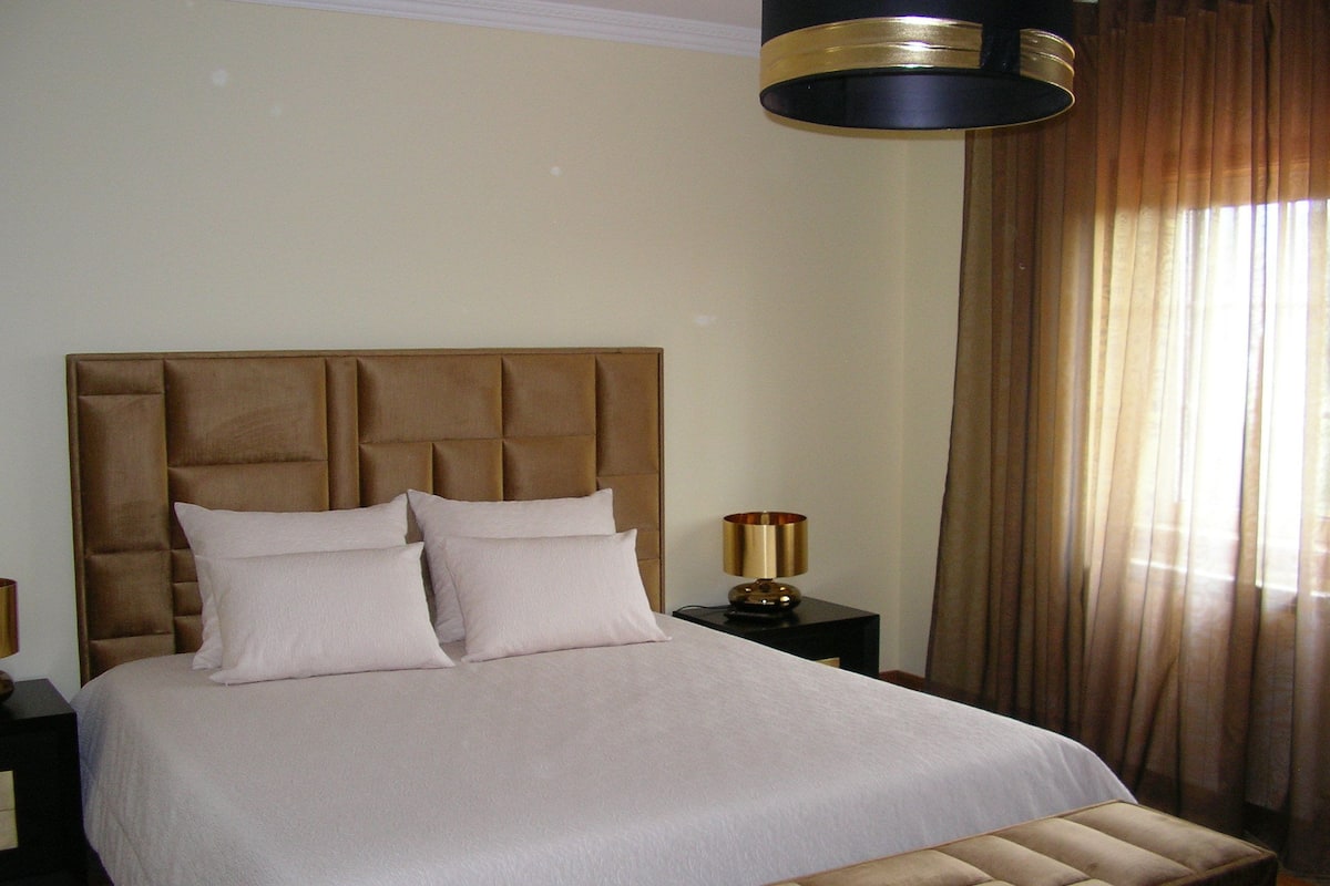 Een net opgemaakt bed met een gestoffeerd hoofdeinde, een bedlamp en een raam met gordijnen.