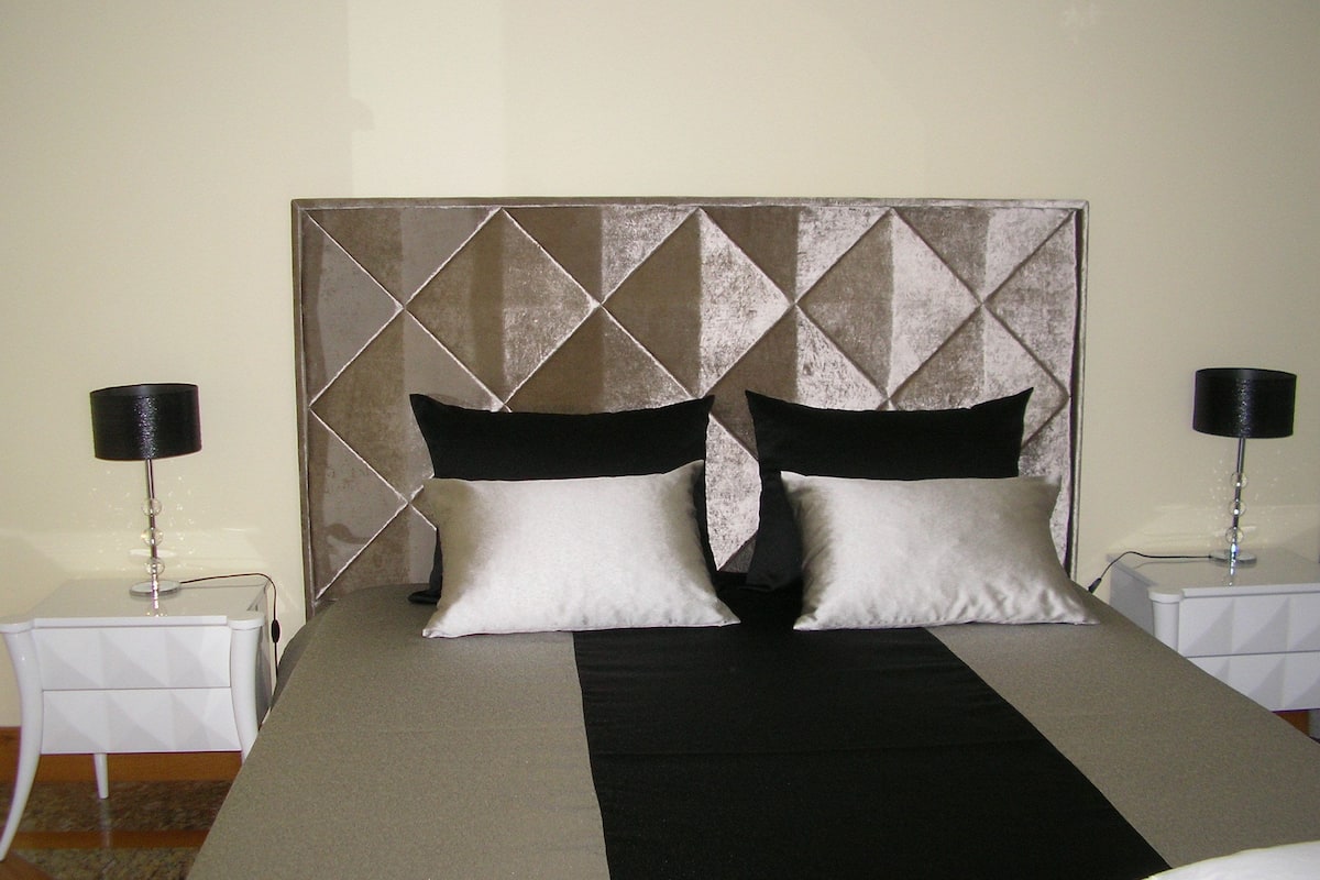 Een keurig opgemaakt bed met een geometrisch hoofdbord, twee nachtkastjes met lampen en een kamerbreed tapijt met dessin.