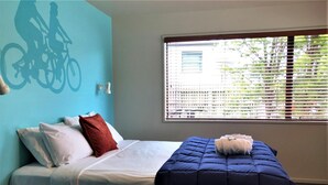 Una cama con ropa de cama blanca y azul, un cojín rojo y una colcha azul. Se ve una ventana con persianas y vistas a follaje verde en el exterior.