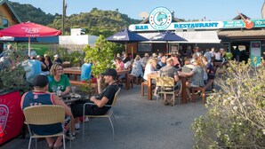 Zona de estar al aire libre en un restaurante frente al mar, con mesas y sillas, rodeada de vegetación y colinas al fondo.