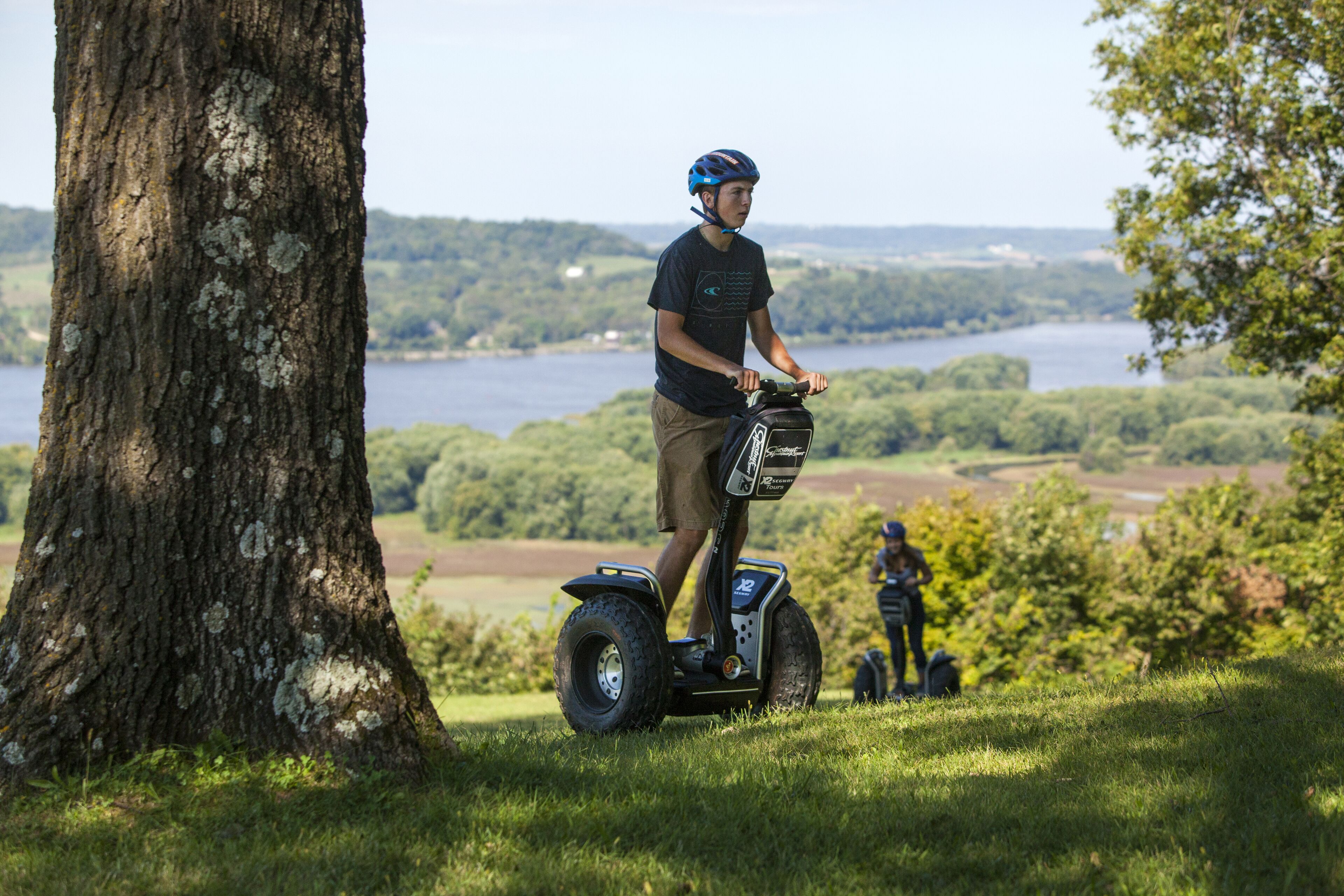 A person riding a Segway on grass near a tree, with a lake and hills in the background.