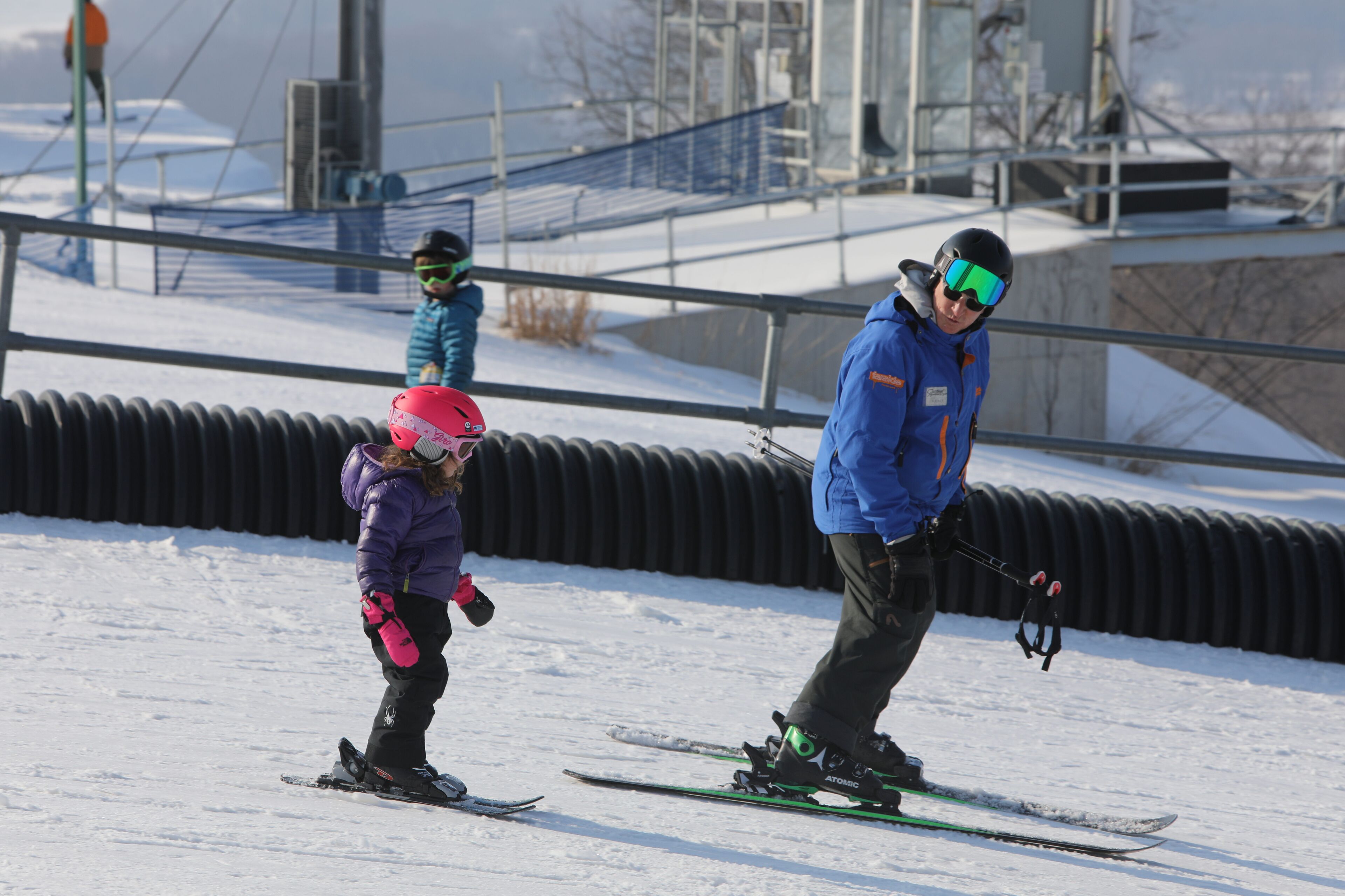 A ski slope with a person skiing and a child in a purple jacket.