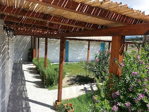 A covered outdoor area with a pergola structure, a pathway leading to a building, and a garden with various plants and flowers. - Sa Corti De Sa Perda (Piscinas)