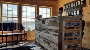 Una habitación rústica con una pared de madera, una estantería llena de libros, un cartel que dice 'RIVERSIDE RANCH' y una ventana con vistas al cielo.