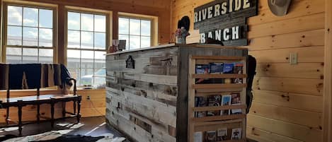 A rustic room with a wooden wall, a bookshelf with books, a sign reading 'RIVERSIDE RANCH', and a window with a view of the sky.