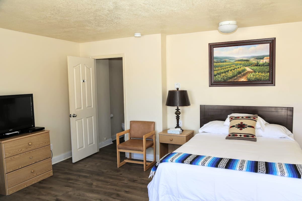 A hotel room with a bed, a TV on a dresser, a chair, a desk, and a painting on the wall.