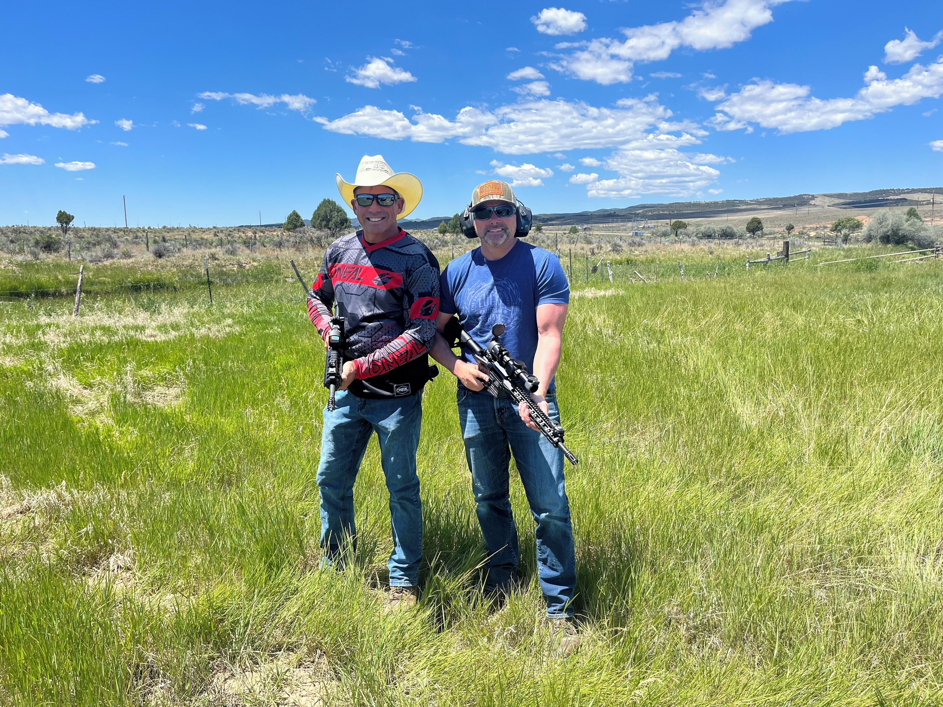 Two people in a field with rifles, wearing casual clothing and cowboy hats.