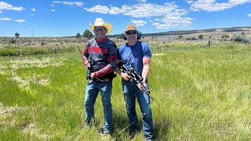 Two people in a field with rifles, wearing casual clothing and cowboy hats.