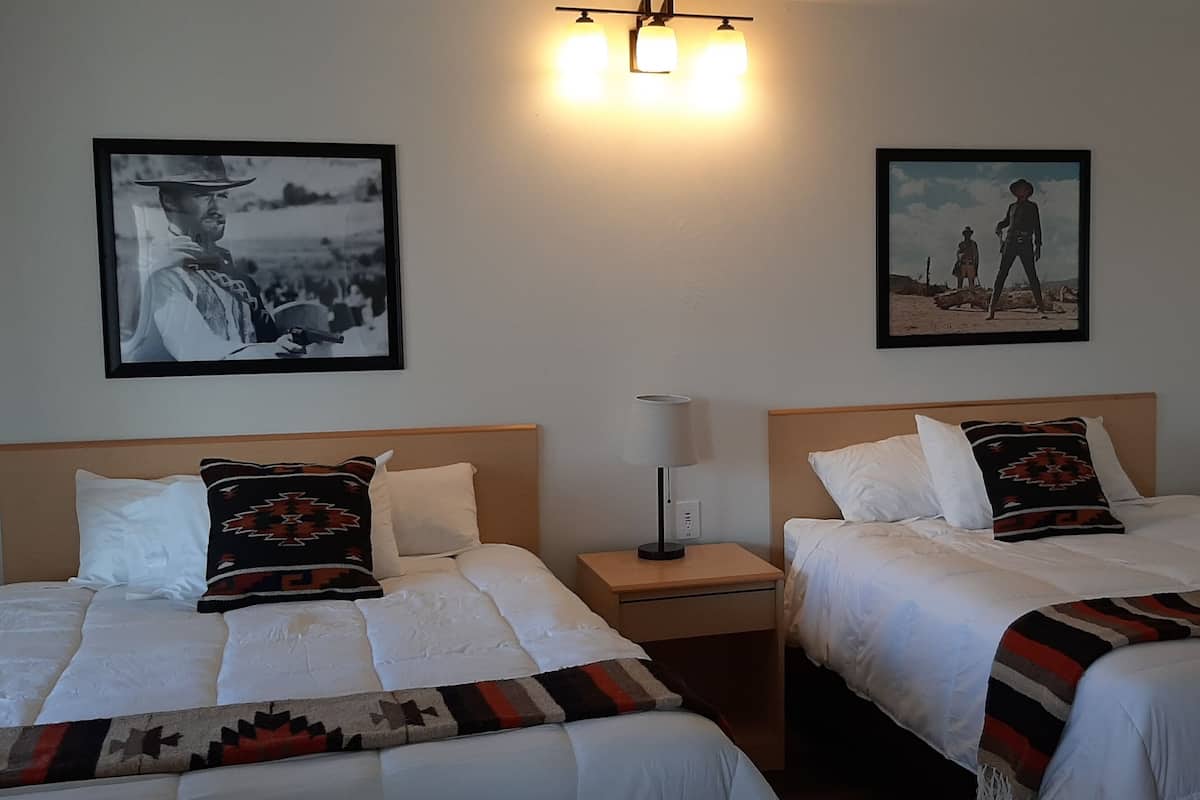 Two single beds with white linens, a wooden headboard, a nightstand with a lamp, and two framed pictures on the wall.