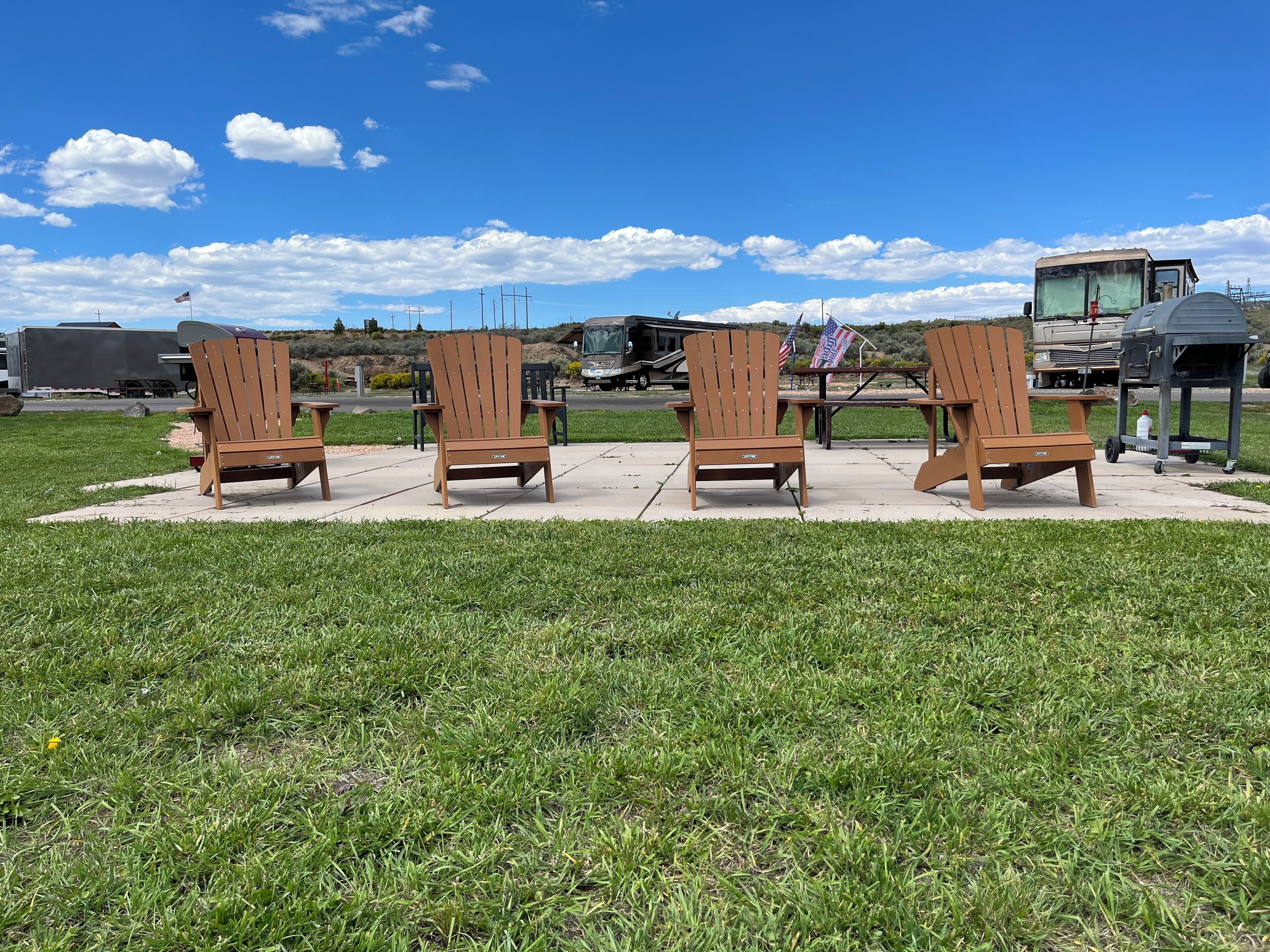 Trois chaises Adirondack en bois sont disposées sur une terrasse en béton, avec un champ verdoyant au premier plan et un ciel bleu dégagé en arrière-plan.