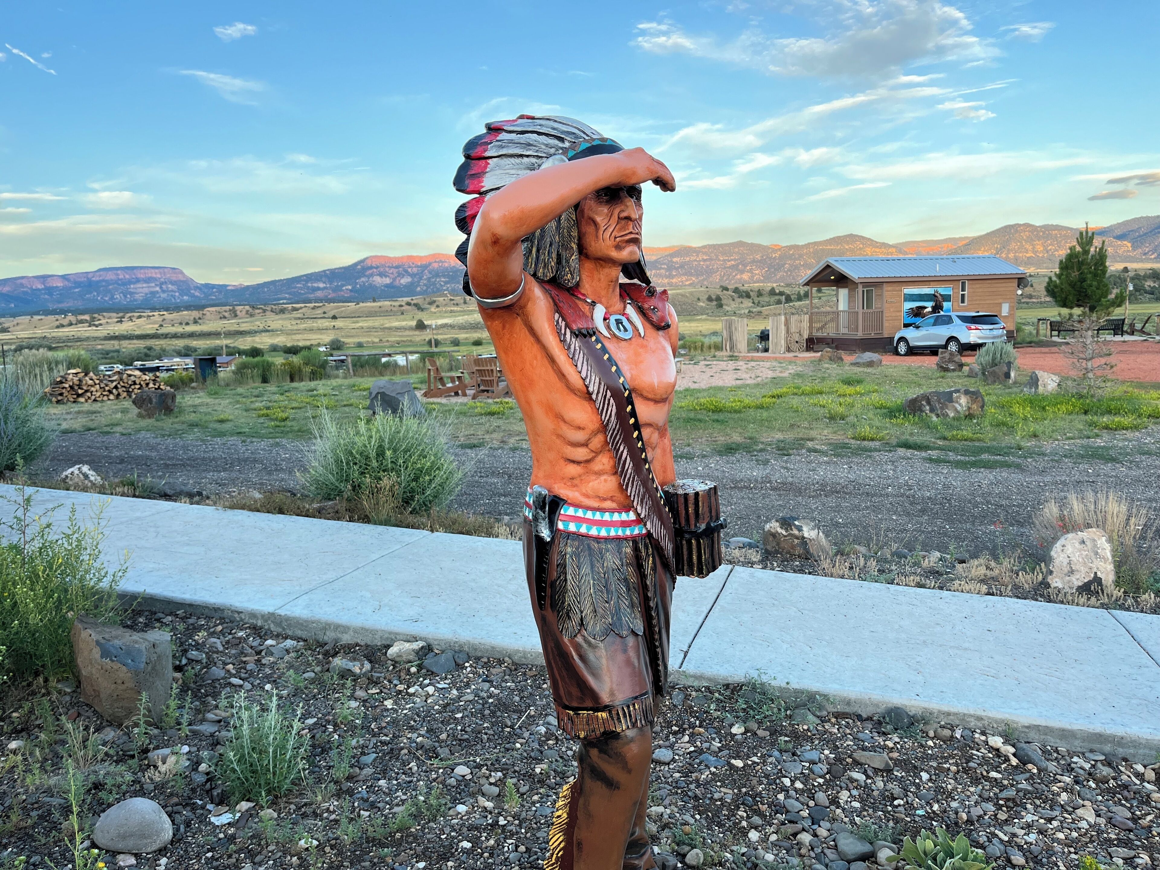A statue of a Native American figure with a feathered headdress, positioned on a concrete ledge overlooking a rural landscape with mountains and a building in the background.