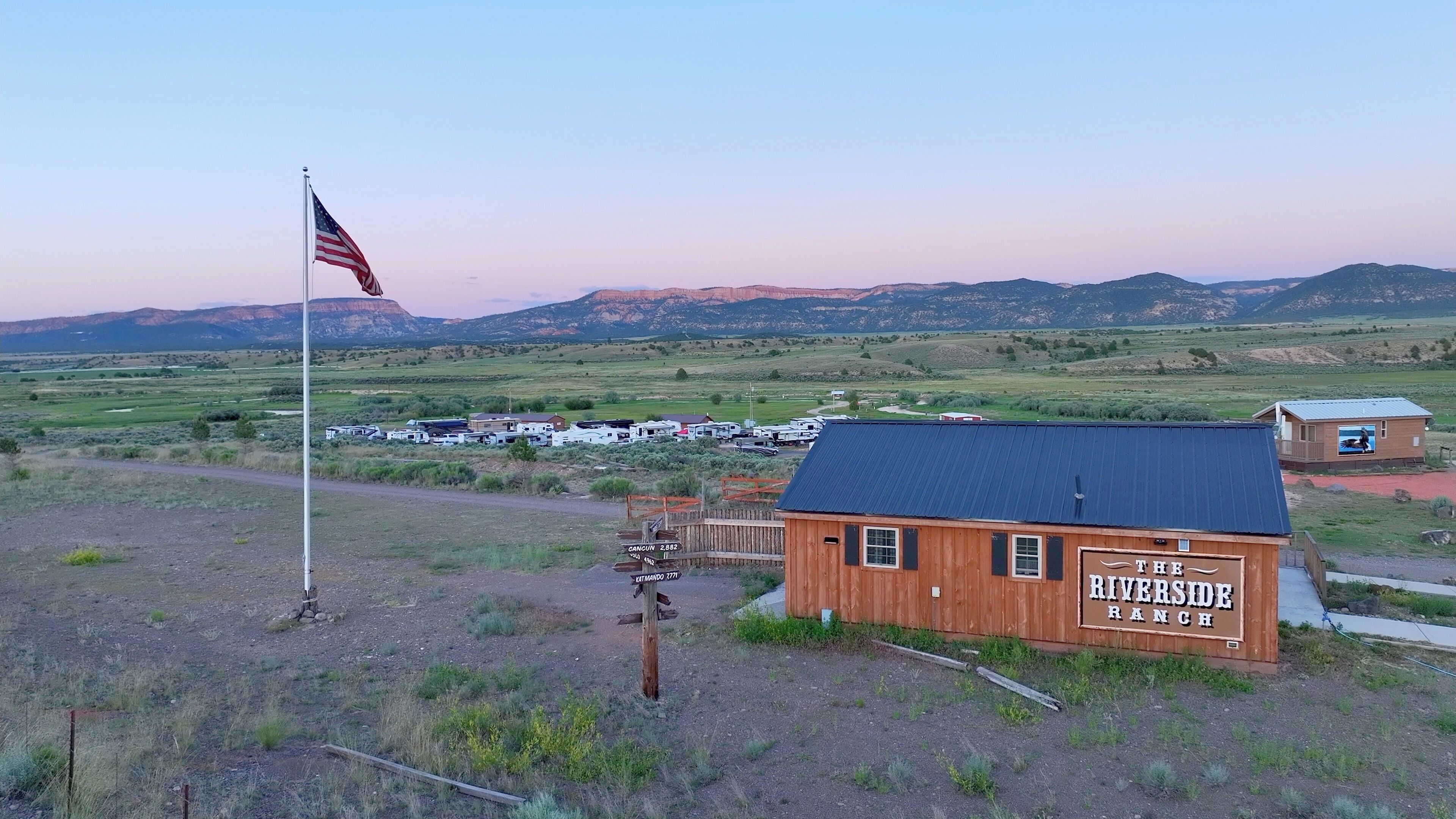 Una construcción de madera con el letrero "THE RIVERSIDE RANCH", un asta de bandera y un cartel en un paisaje rural.