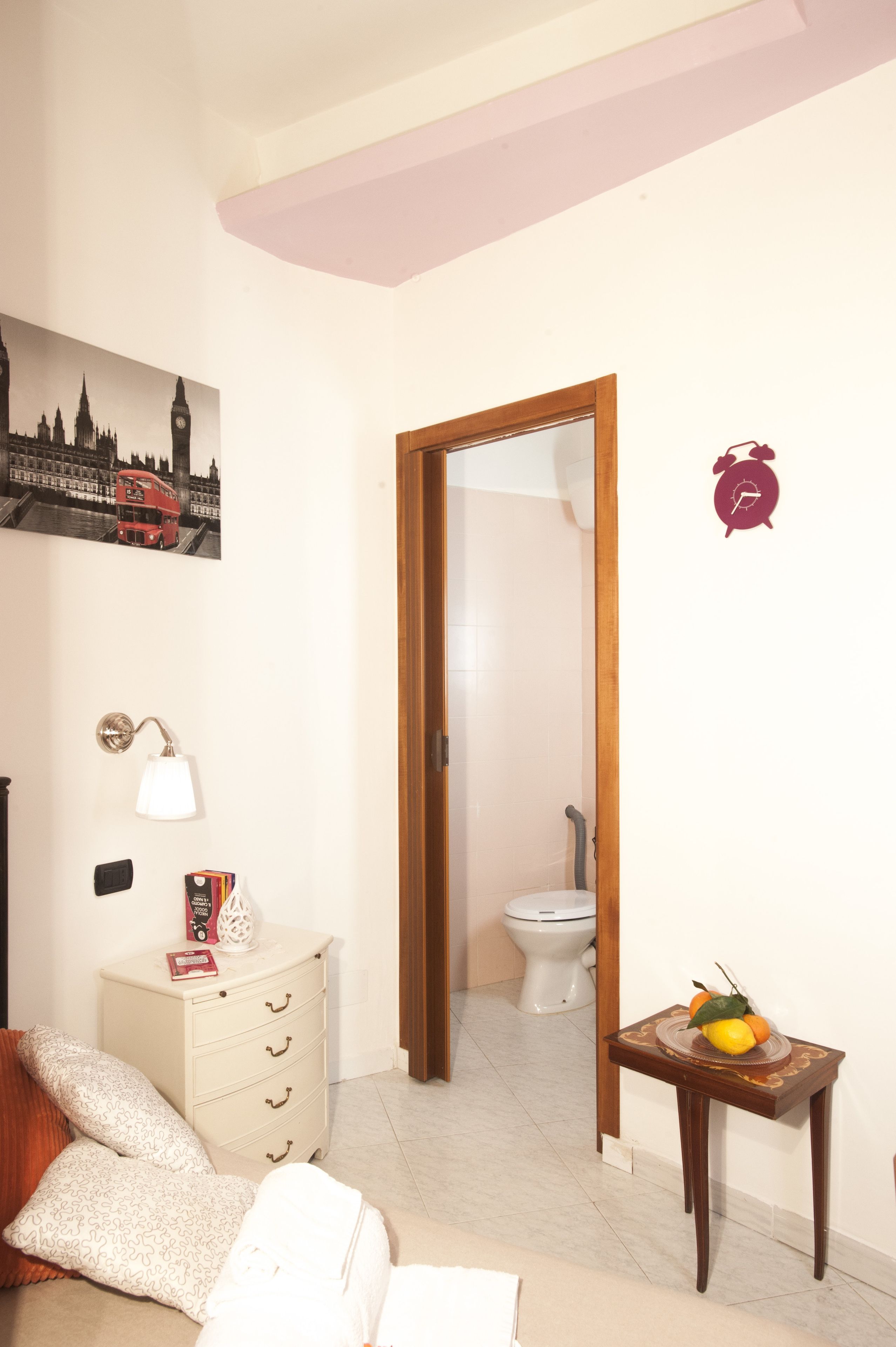 Una habitación con una cama, un aparador, una lámpara, un cuadro de los monumentos de Londres, una puerta de madera y una pequeña mesa con fruta.
