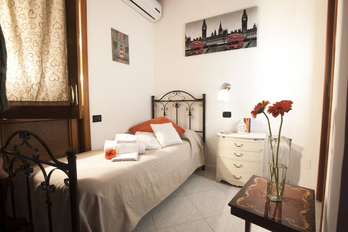 Una cama bien tendida con cabecera, una mesita de noche con un florero y una pared con un cuadro de paisaje urbano.