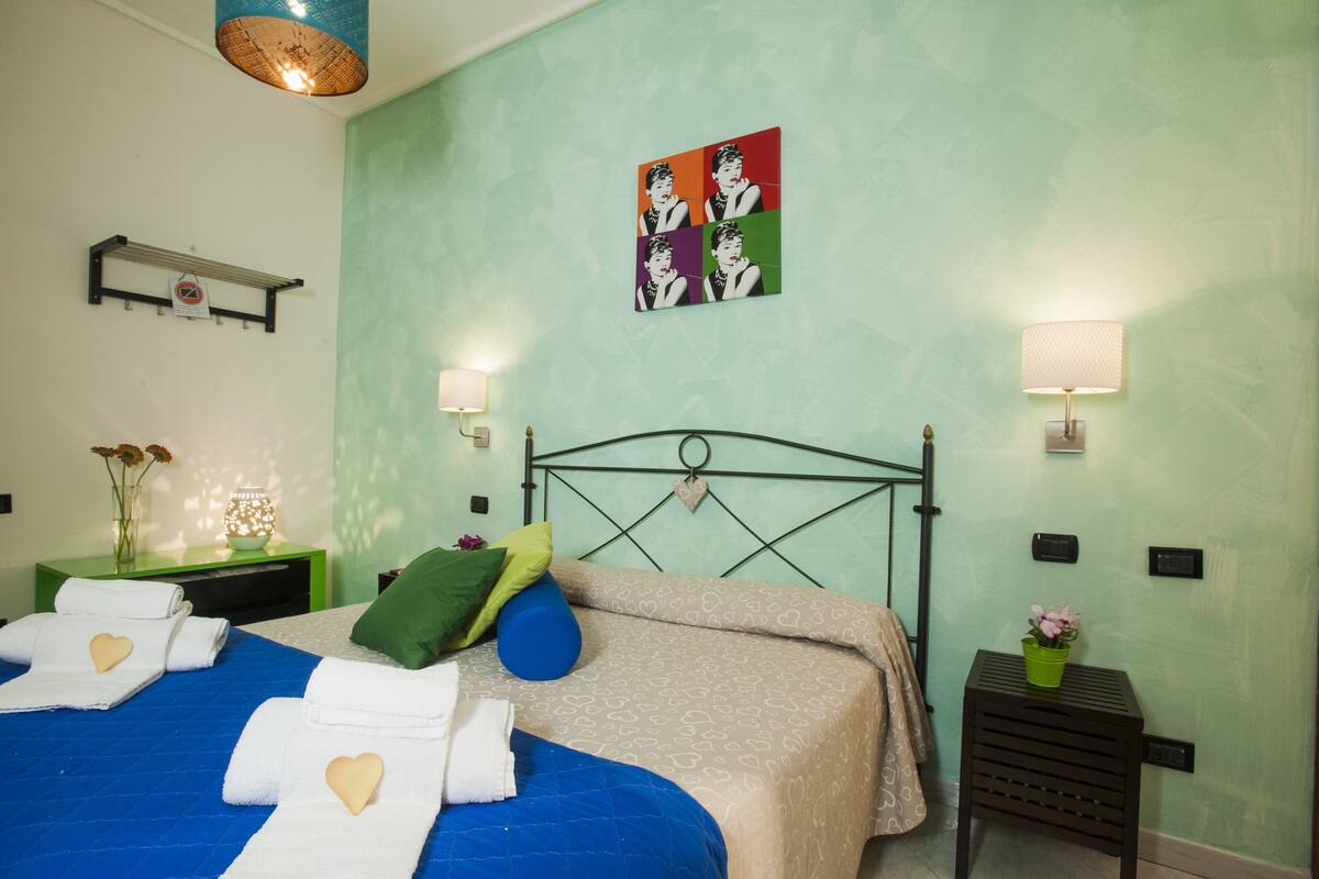 Una cama bien hecha con toallas en forma de corazón, una mesita de noche verde con una lámpara, una obra de arte en la pared y una pequeña planta en maceta.