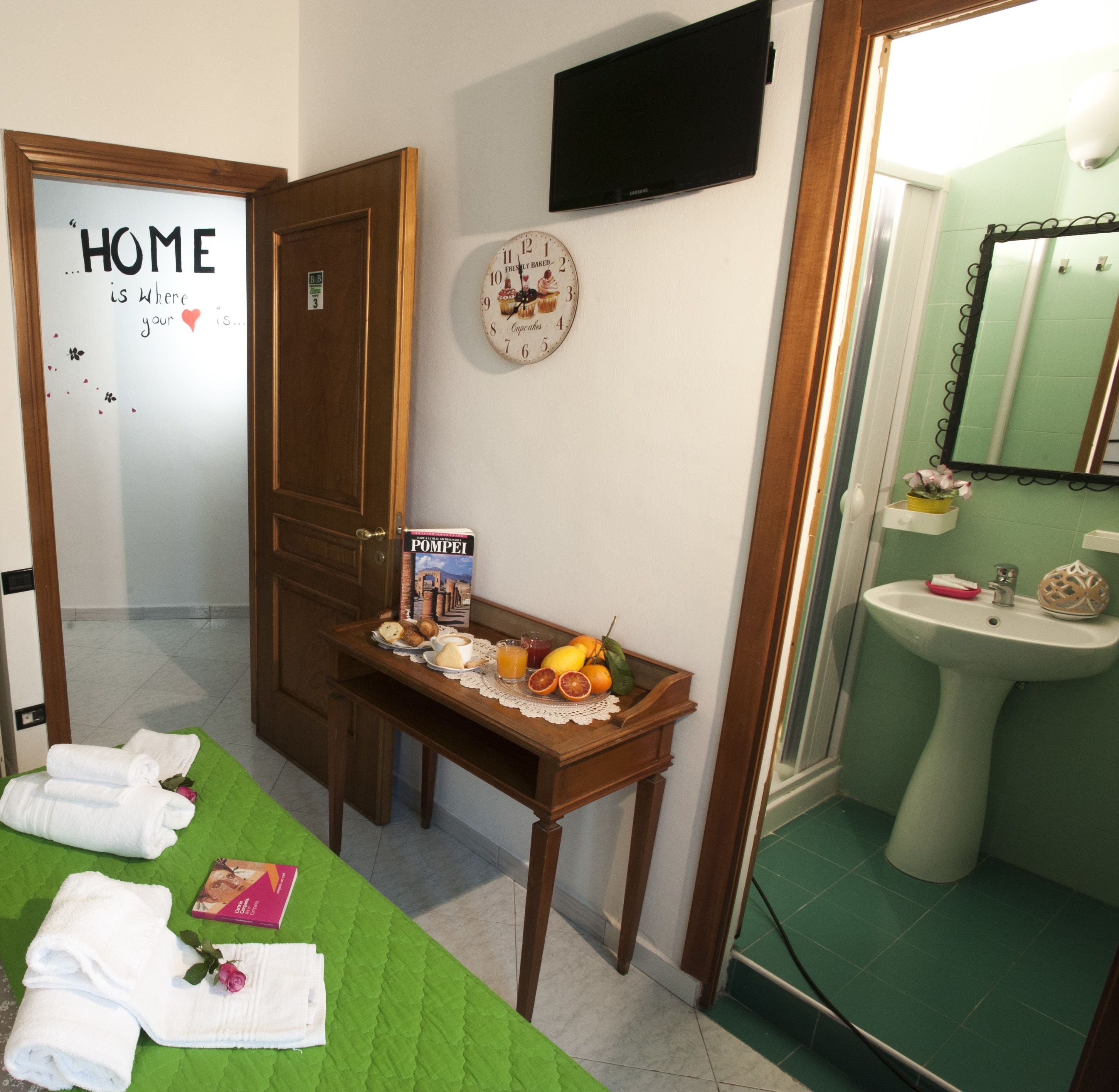 A hotel room with a green bedspread, a table with a book and fruit, a clock with cupcake illustrations, and a bathroom with a sink and mirror.