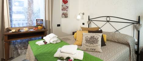 Una cama bien hecha con un edredón verde, una mesita de noche con un cartel que dice "POMPEI" y una ventana con vistas a edificios.