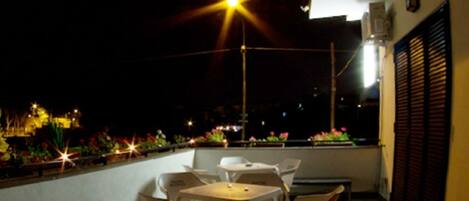 An outdoor patio with tables and chairs, a street view with lights, and a building with shutters.