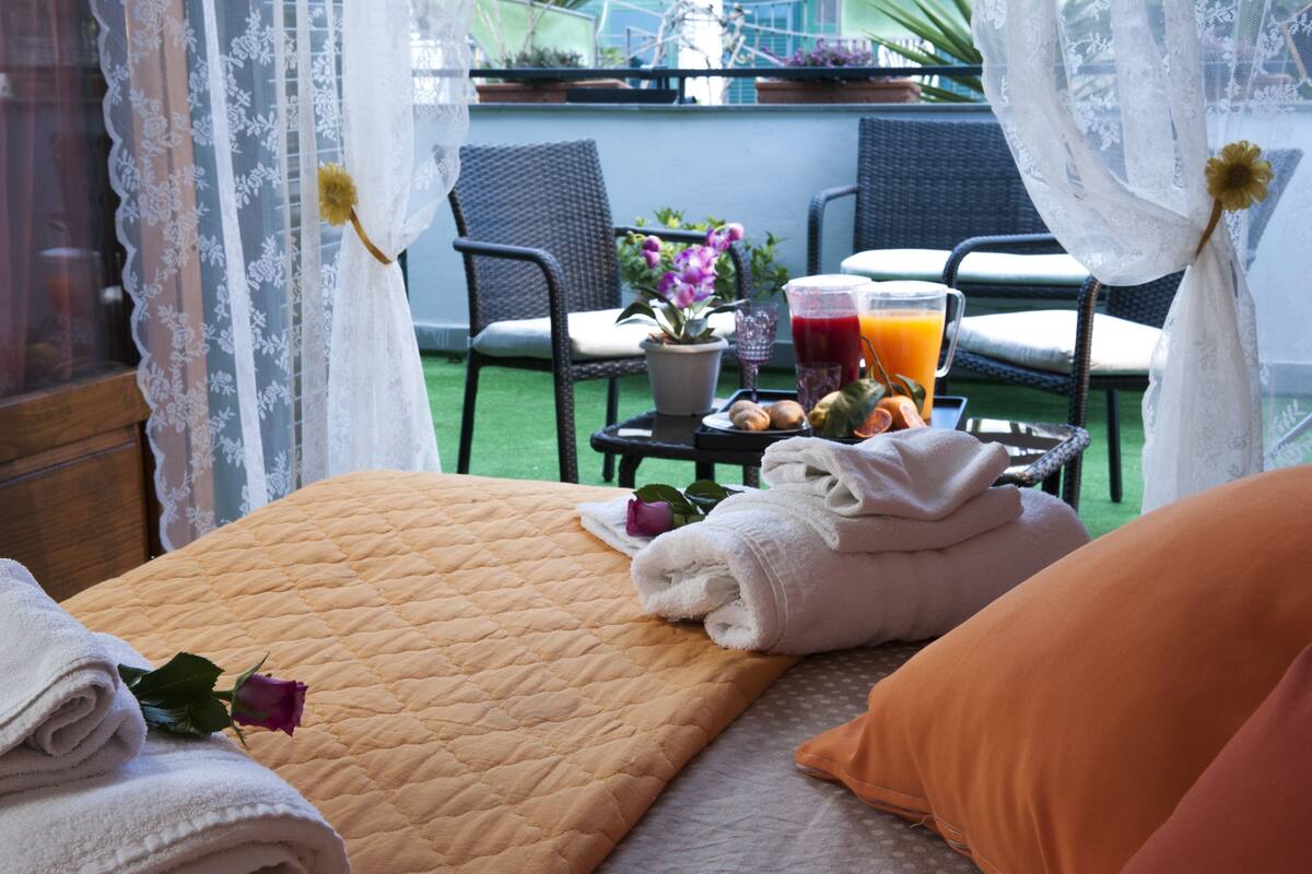 Habitación de hotel con una cama, una mesita con una cesta de frutas, un vaso de zumo de naranja y una planta en maceta.