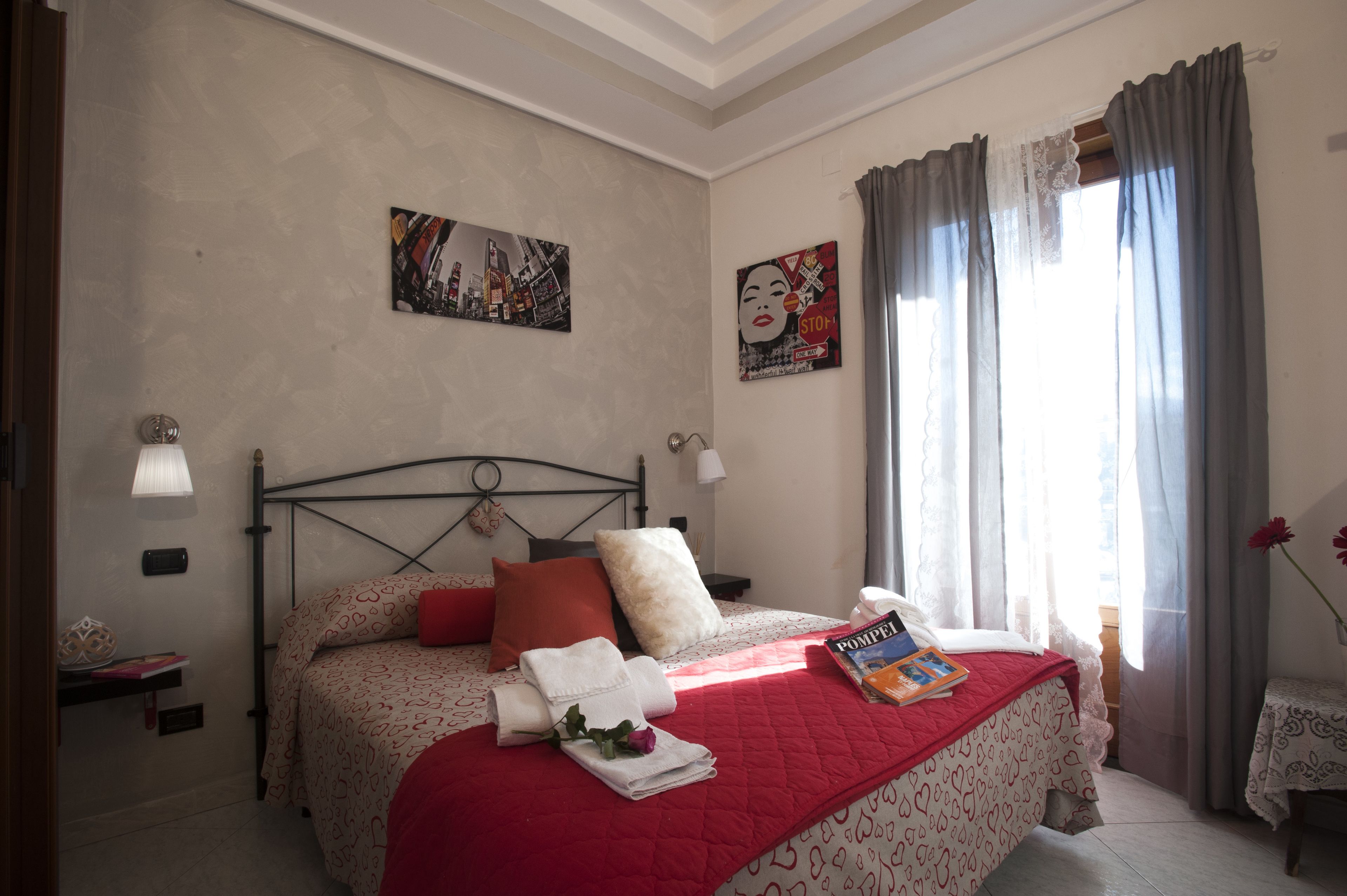 A neatly made bed with a red blanket, pillows, and a book. There are two wall-mounted lamps, a painting, and a window with curtains.