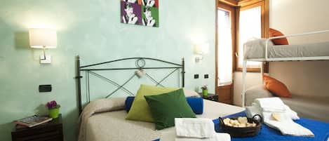A neatly made bed with a blue quilt, green and blue pillows, and a heart-shaped towel. There is a small table with a plant and a magazine. A colorful artwork is on the wall, and a window with curtains is visible.