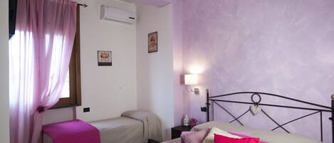 Quarto com duas camas de solteiro, janela com cortinas cor-de-rosa, ar-condicionado fixado na parede, luminária de cabeceira e um objeto decorativo na parede.