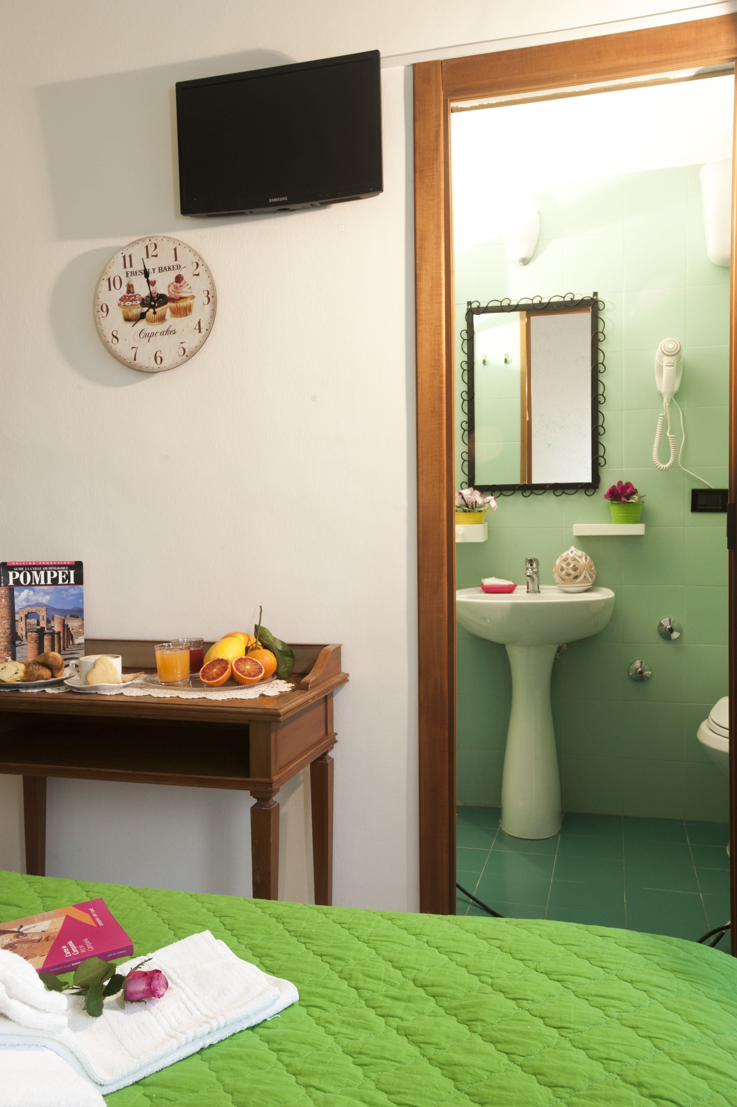 Una habitación con una cama, una mesita de noche con un libro, un reloj, un lavabo y un espejo.