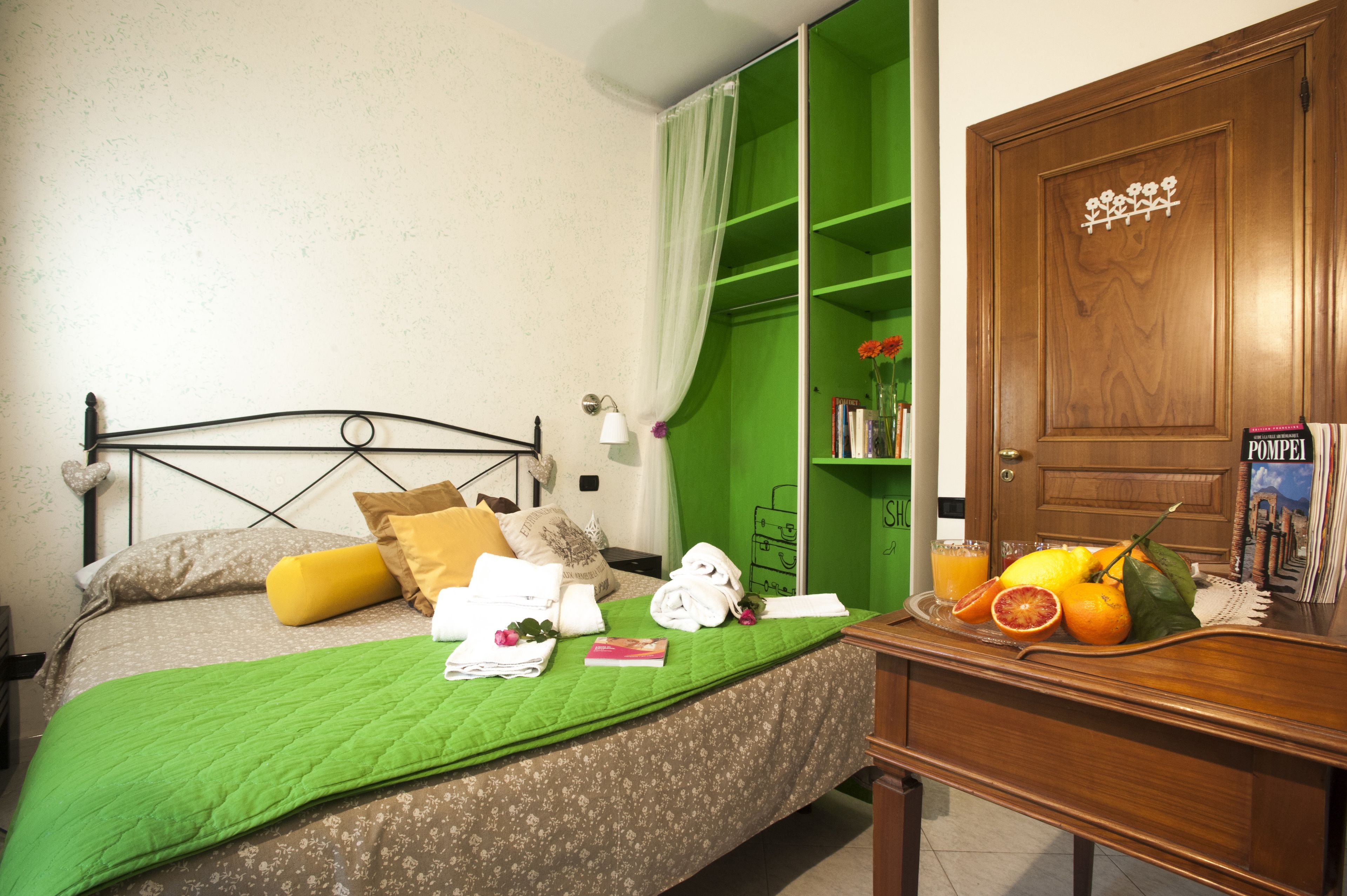 Quarto com cama, cortinas verdes, guarda-roupa de madeira e mesa de cabeceira com frutas e um livro.
