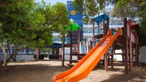 A playground with a slide, surrounded by trees and a building with a blue and white facade. - Mariant (Sant Llorenc des Cardassar)