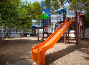 A playground with a slide, surrounded by trees and a building with a blue and white facade. - Mariant (Sant Llorenc des Cardassar)