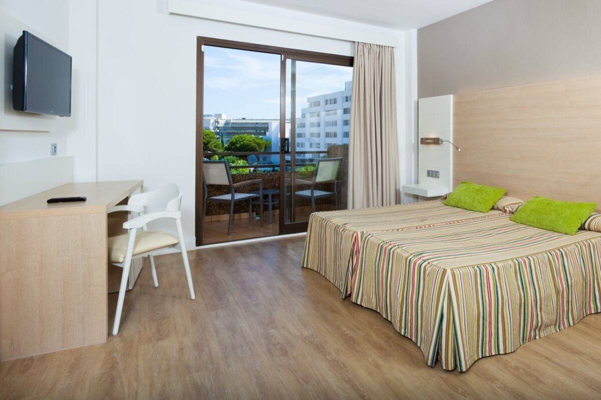 A modern hotel room with a bed, a desk, a chair, a TV, and a balcony with a view of buildings and greenery.