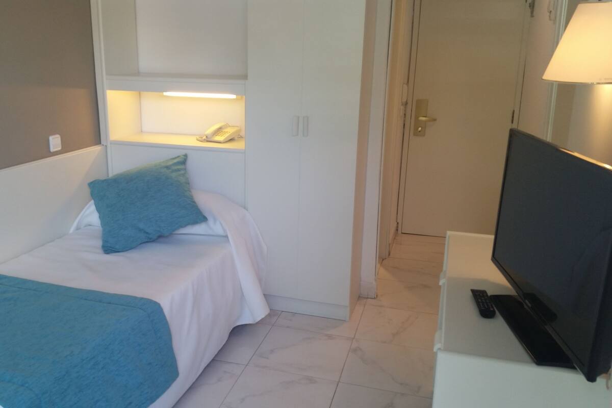 A small, compact room with a single bed, a television on a stand, and a door leading to another room.