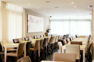 Un salón de conferencias con mesas y sillas largas, una pantalla de proyector y un amplio ventanal con persianas.
