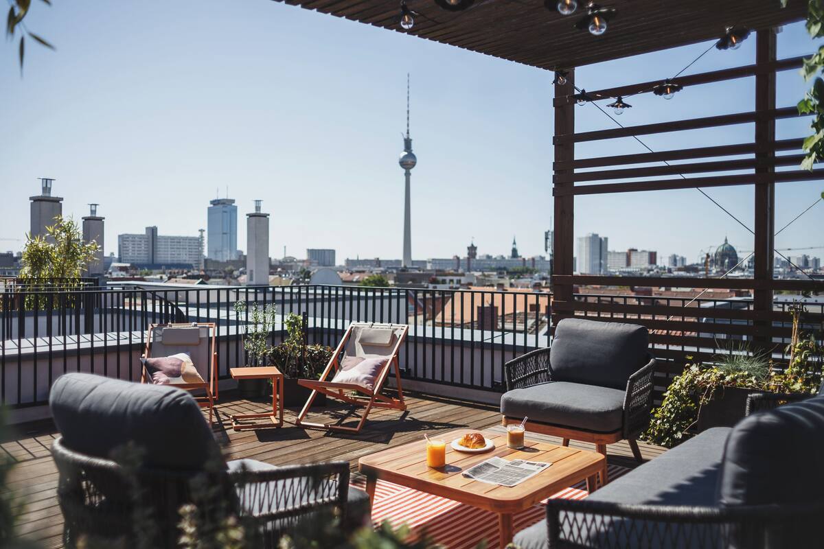Eine Dachterrasse mit Sitzgelegenheiten im Freien, einem Holztisch und Blick auf die Stadtlandschaft, darunter ein markanter Turm.