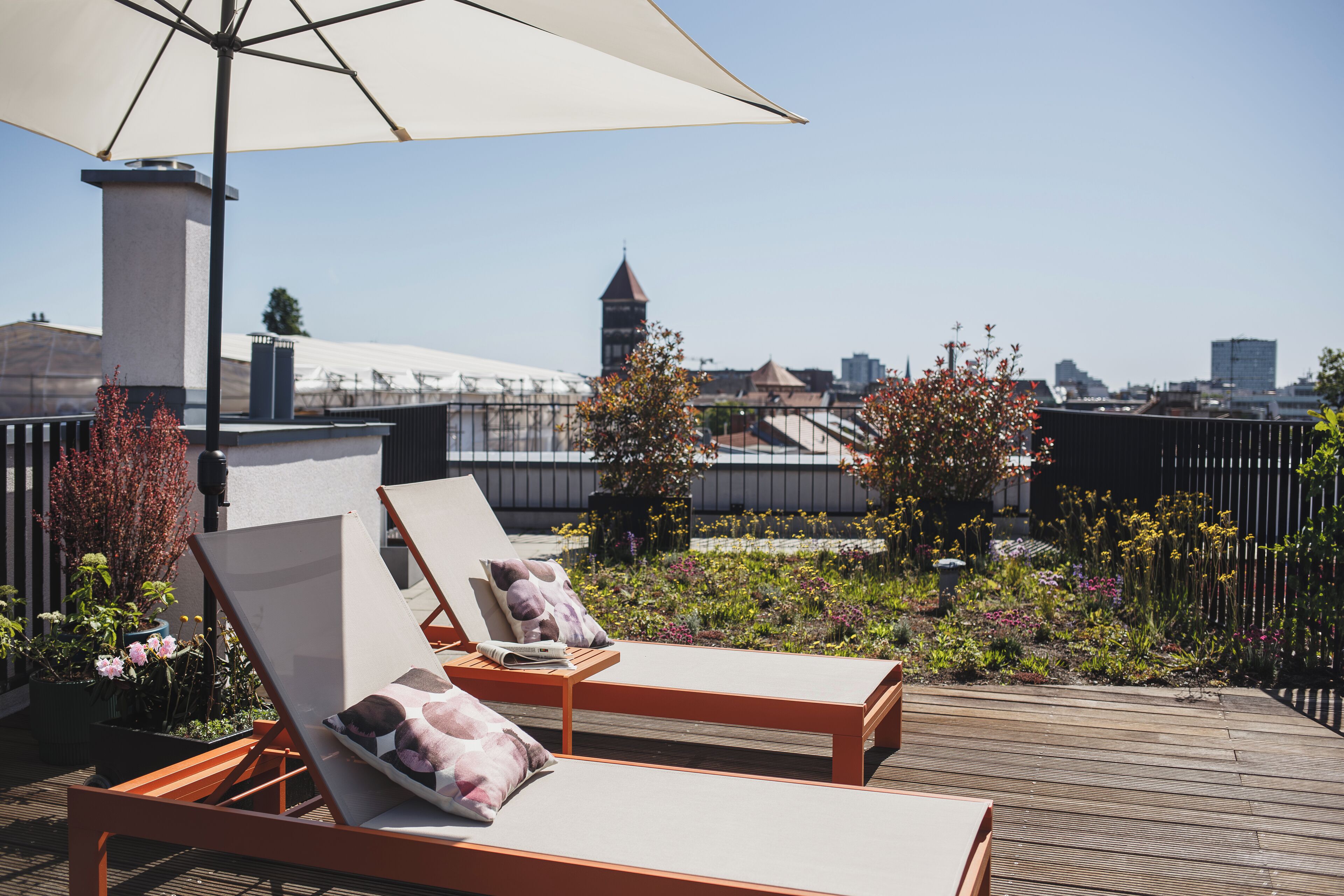 A rooftop terrace with two lounge chairs and a large umbrella, surrounded by potted plants and flowers.