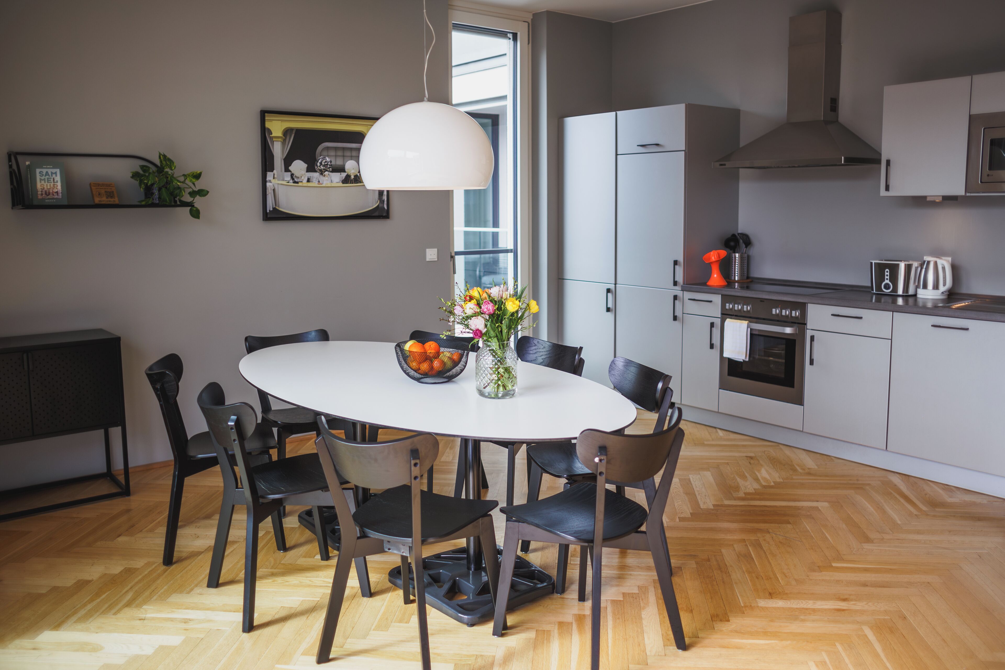 A modern kitchen with a dining area, featuring a white table, black chairs, and a herringbone wood floor.