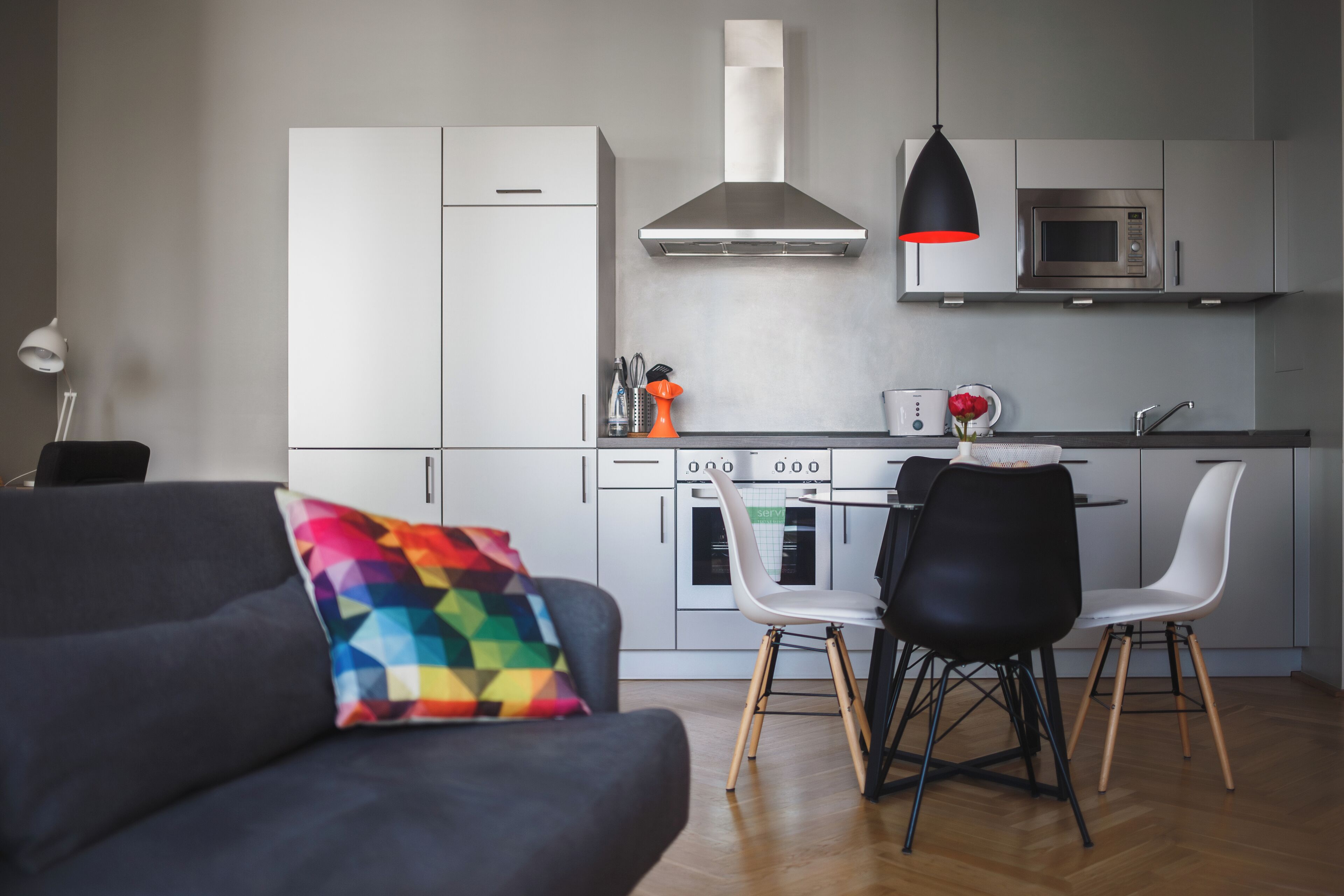A modern kitchen with white cabinets, a stainless steel range hood, and a microwave. There are two black chairs and a colorful geometric pillow on a dark sofa.