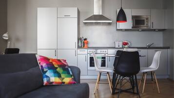 A modern kitchen with white cabinets, a stainless steel range hood, and a microwave. There are two black chairs and a colorful geometric pillow on a dark sofa.