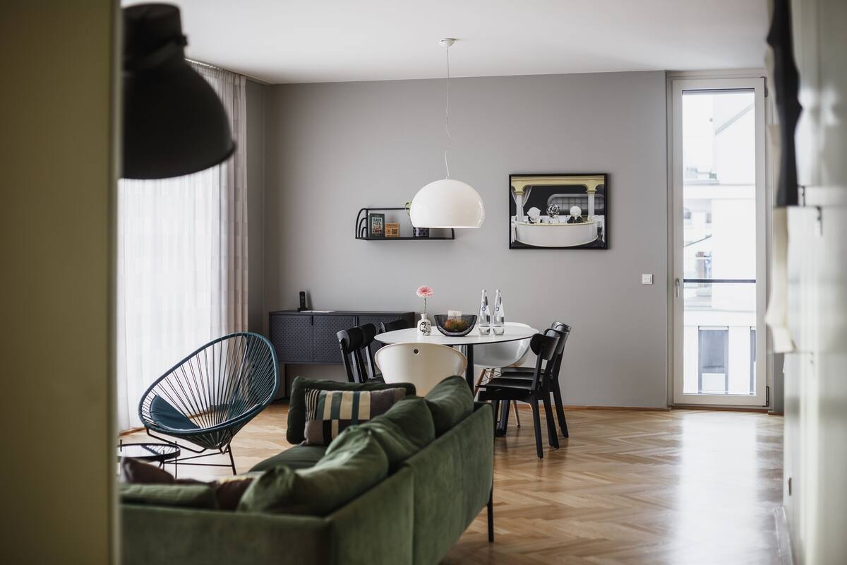 Ein modernes Wohnzimmer mit einem grünen Sofa, einem Esstisch und Stühlen. Im Hintergrund ist eine Küchenzeile mit einer weißen Pendelleuchte und einem gerahmten Bild an der Wand zu sehen.