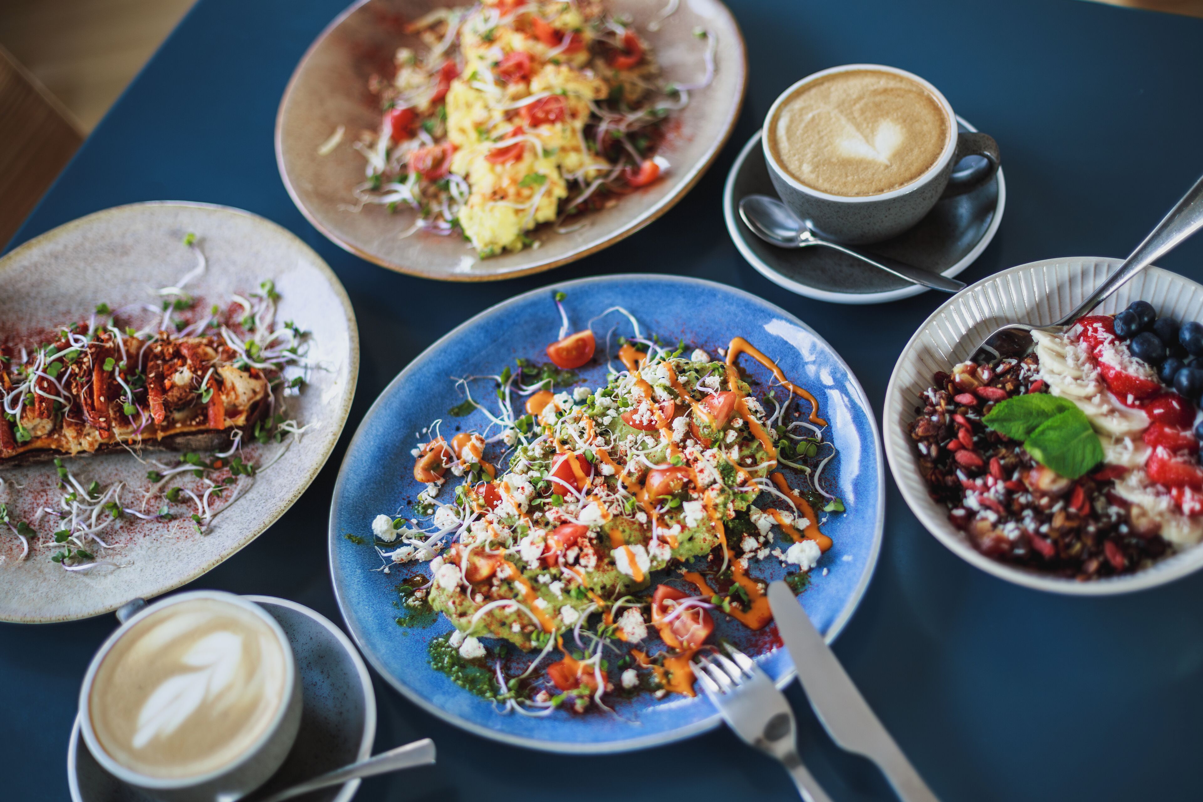 A table with various dishes, including a blue plate with a salad, a plate with grilled fish, and a bowl of berries. There are also two cups of coffee.