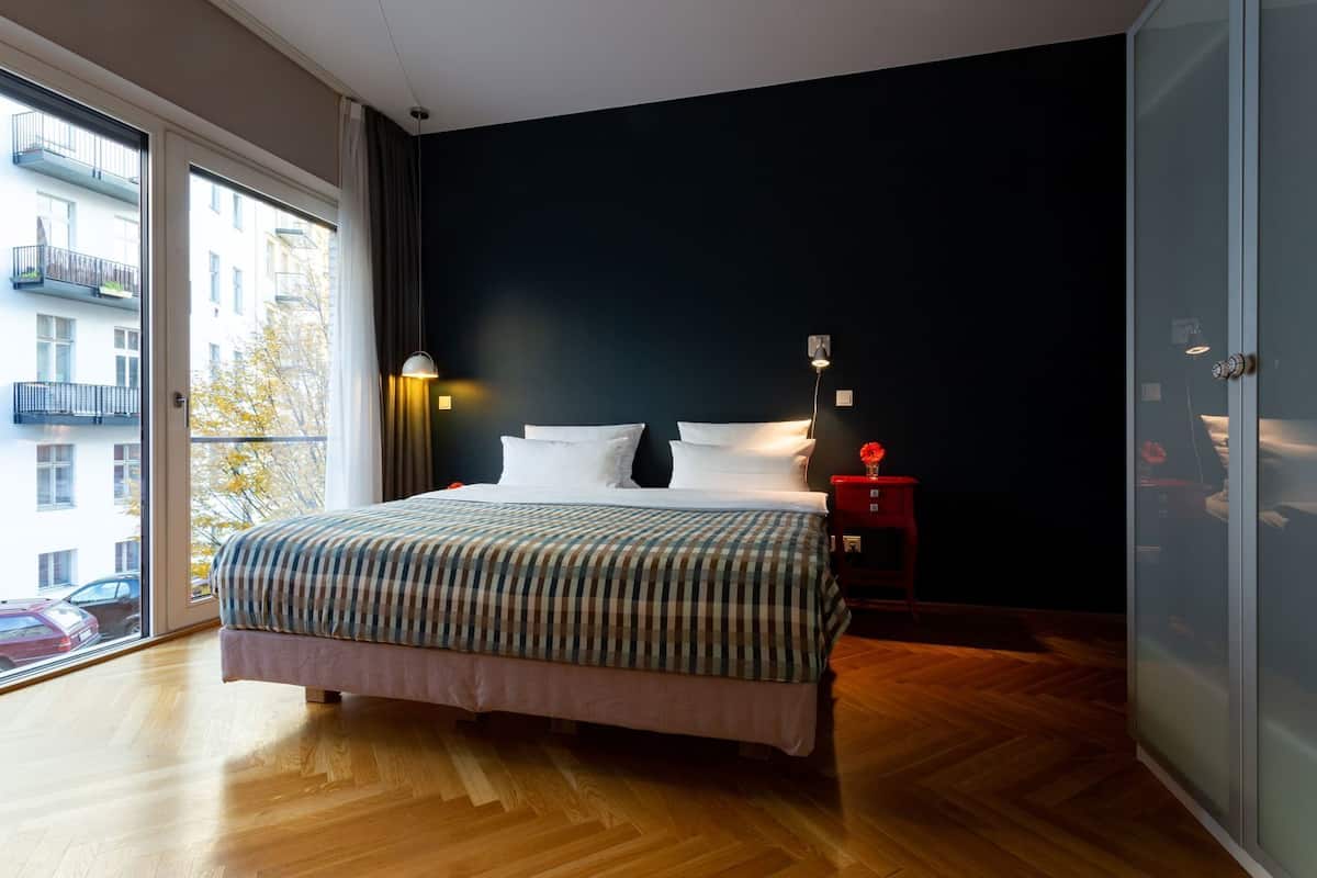 Ein Schlafzimmer mit einem großen Bett, einem Nachttisch mit Lampe, einem Fenster mit Vorhängen und Blick auf ein Gebäude.