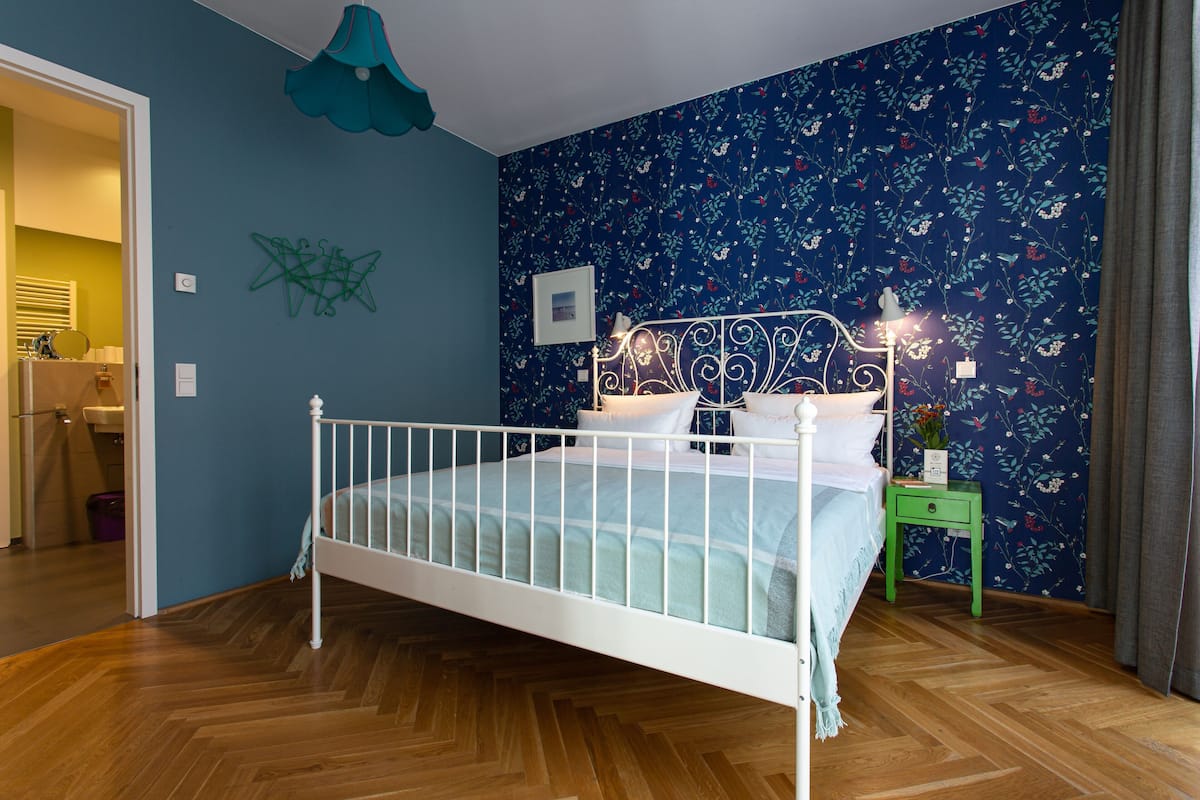 Ein Zimmer mit einem weißen Metallbett, einem grünen Nachttisch, einer blauen Wand mit Blumenmuster und einem Holzboden im Fischgrätmuster.
