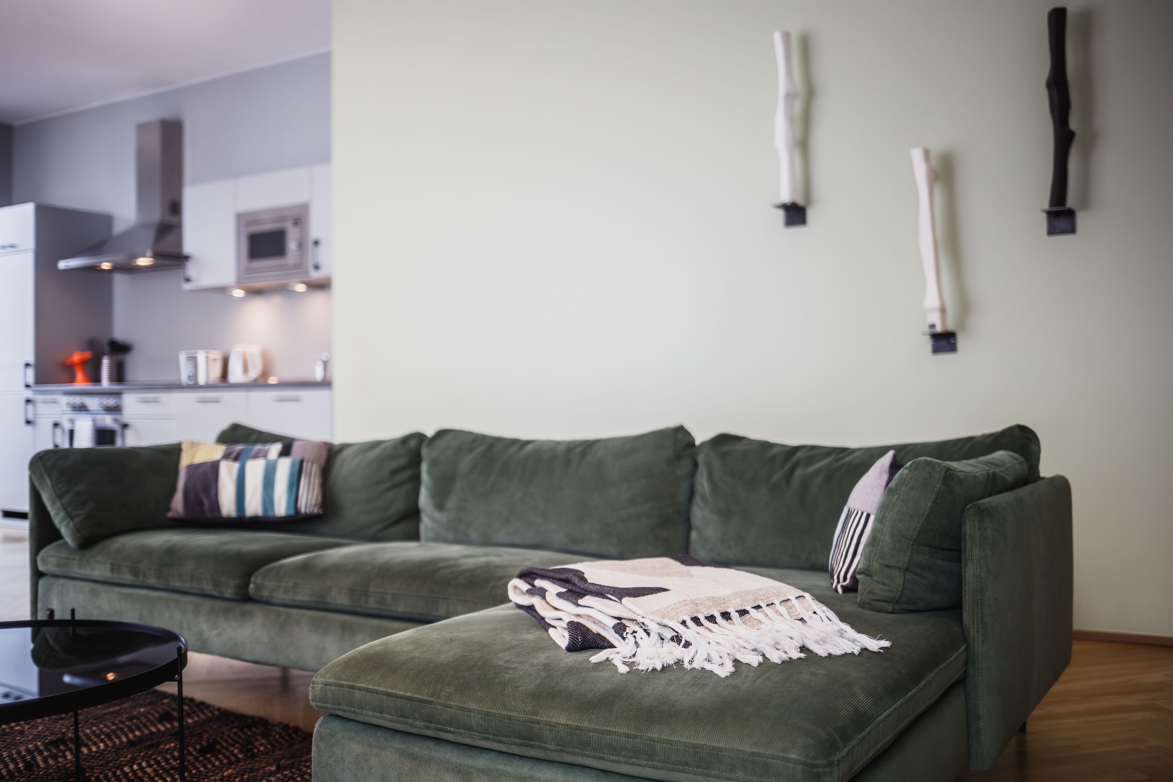Un sofá verde con una manta, una cocina al fondo y una pared con tres objetos verticales.