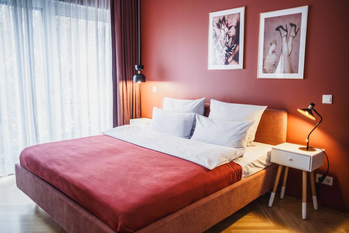 Ein ordentlich bezogenes Bett mit weißen Leinen, einer roten Tagesdecke, einem Nachttisch mit Lampe und zwei gerahmten Kunstwerken an der Wand.