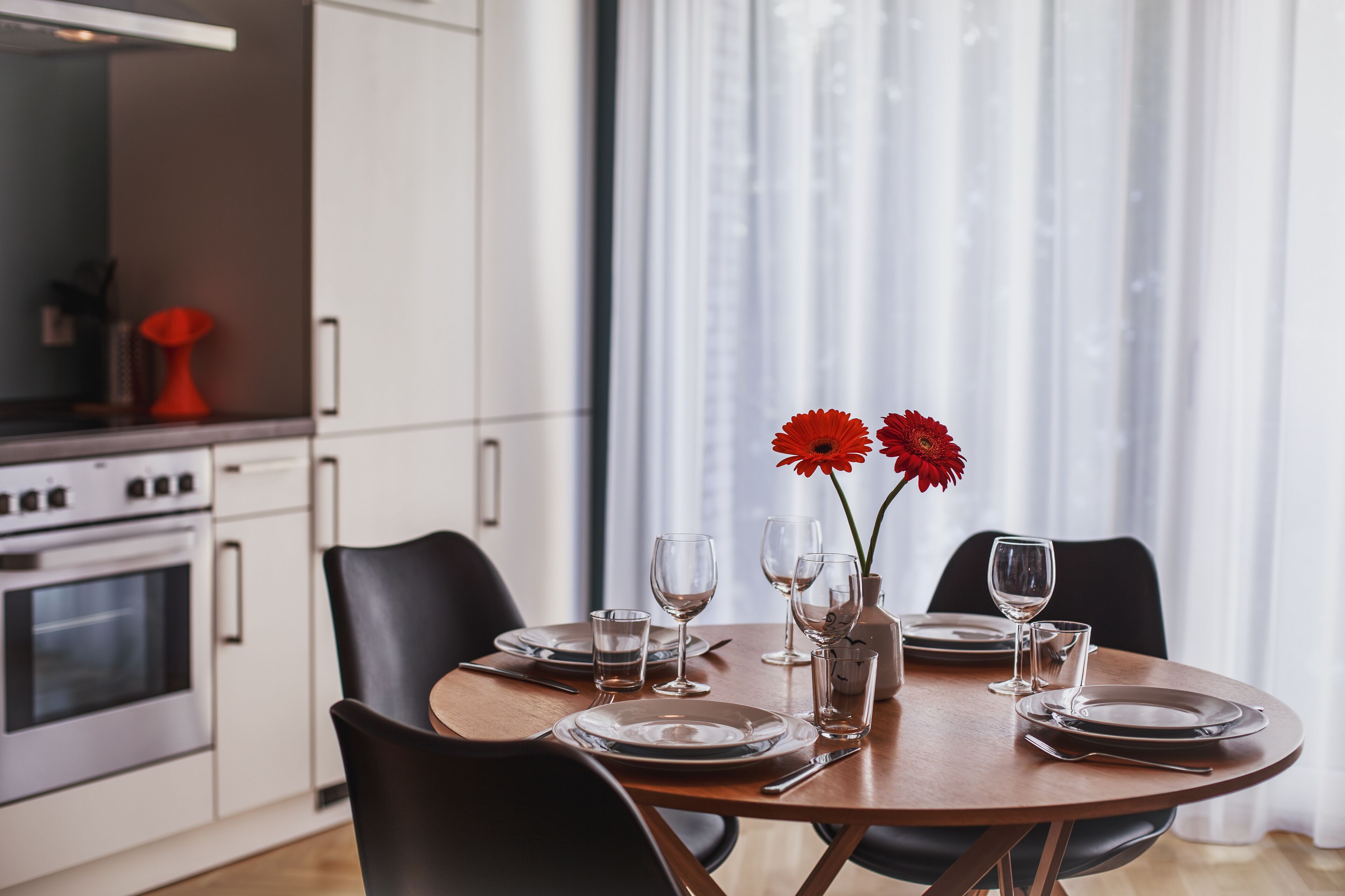 A dining table set with plates, glasses, and a vase with red flowers, in a modern kitchen with white cabinets and a large window.